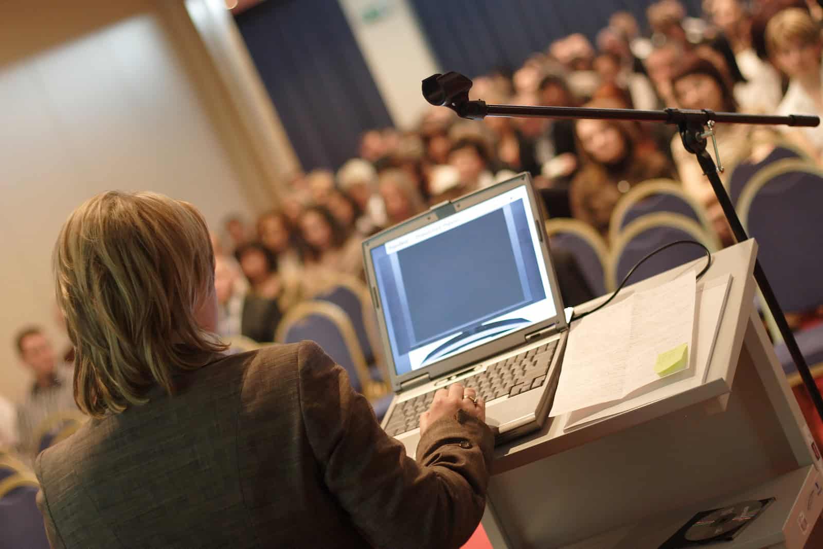 Business woman at podium with laptop computer lecturing audience in auditorium